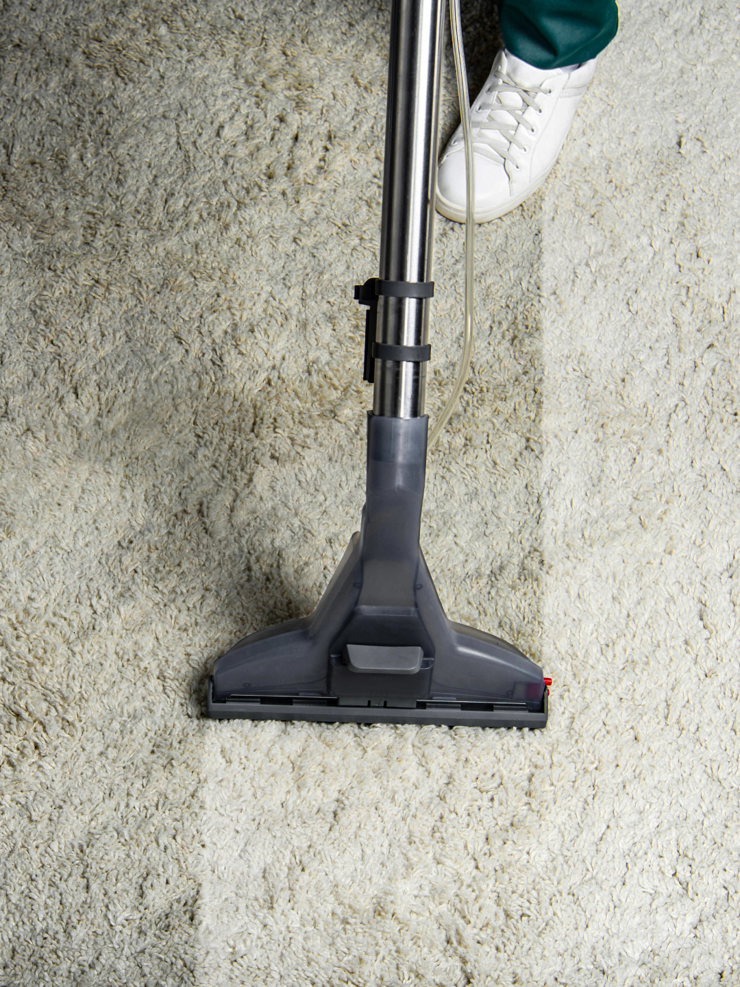 A vacuum cleaner head positioned on a light-colored carpet, with a shoe wearing a white sneaker visible in the lower corner.
